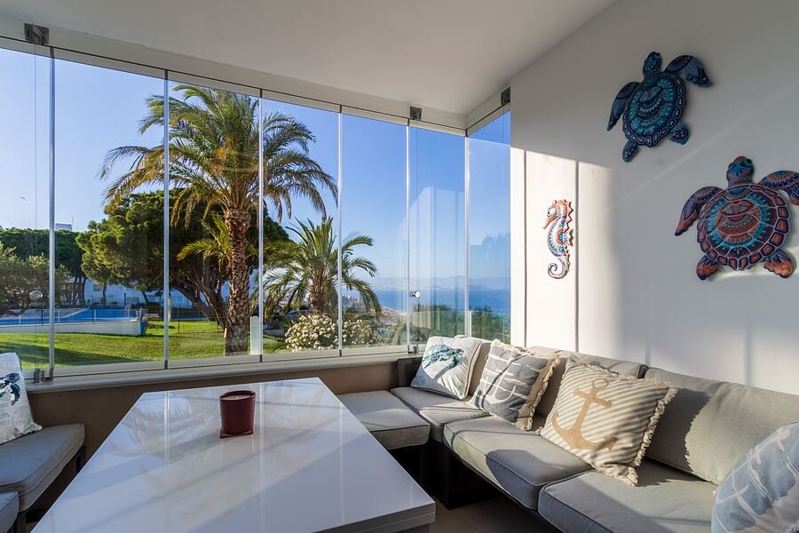 Apartamento Gran Alacant: galería acristalada con sofá rinconero, palmeras y el mar al otro lado del cristal