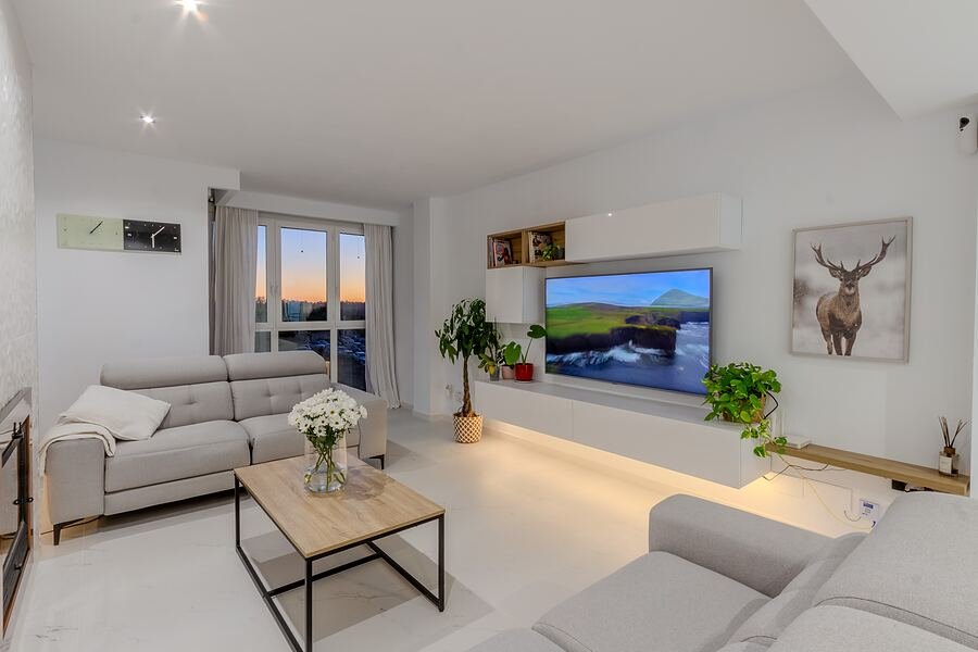 Ático El Altet: salón alargado en blanco con sofá gris, televisor en mueble bajo y ventanal al atardecer