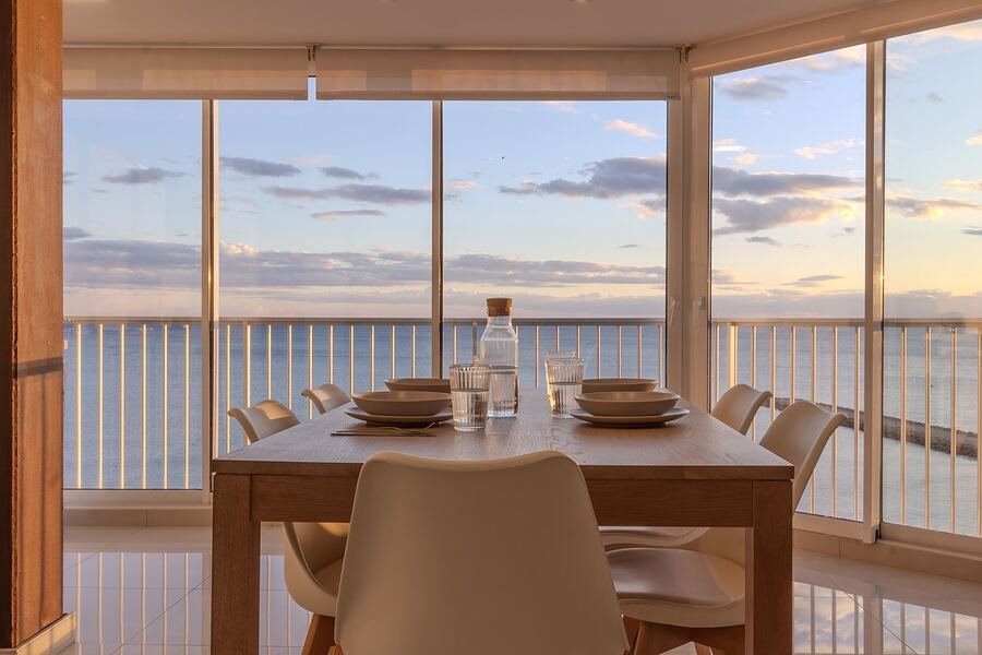 Ático Levante en Santa Pola: comedor de madera frente al ventanal corrido, con el mar y el cielo del atardecer al fondo